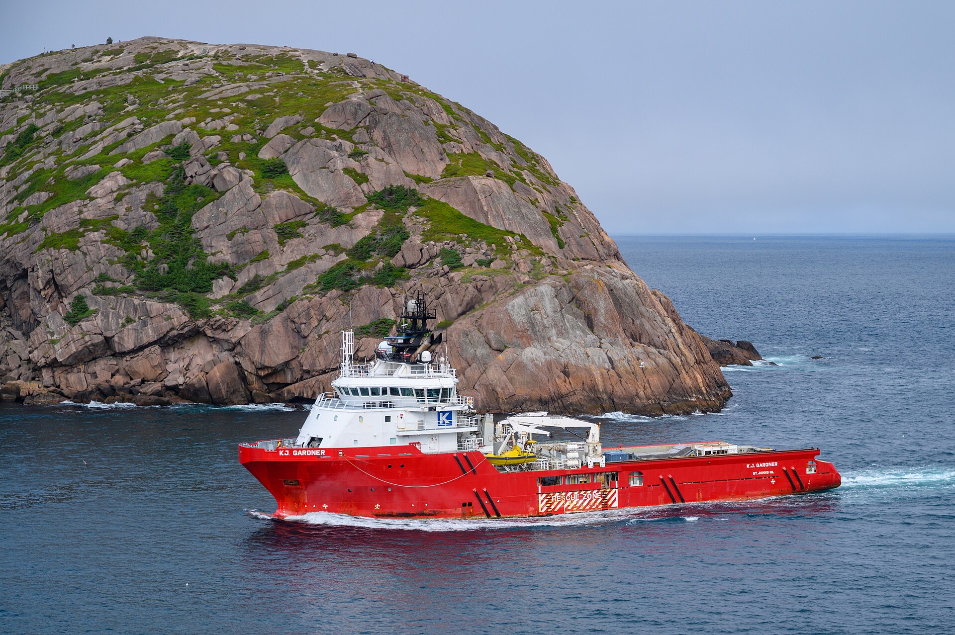 Navio de apoio offshore vermelho navegando junto à costa rochosa